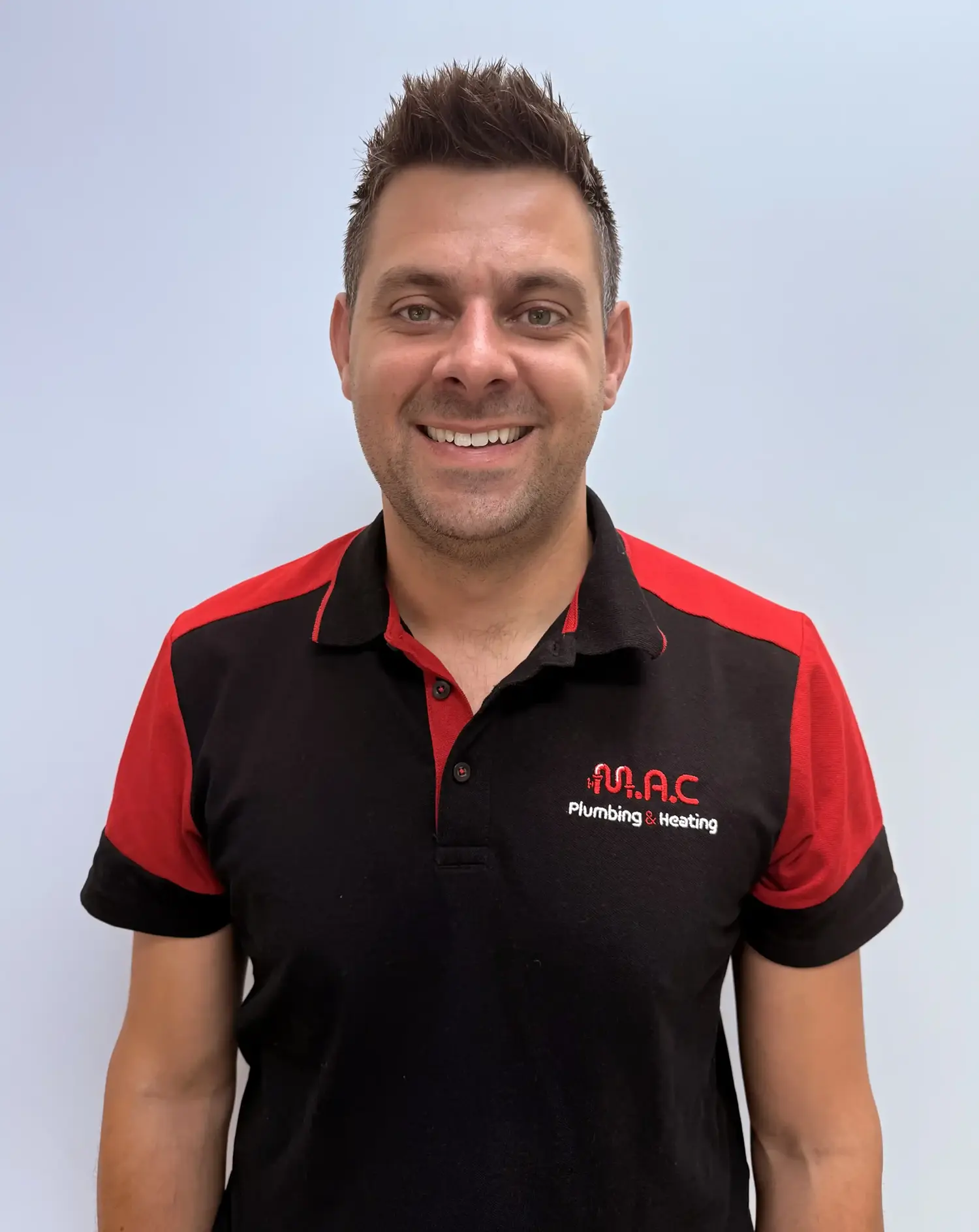 Man wearing a black and red polo shirt with M.A.C Plumbing & Heating logo, smiling at the camera.